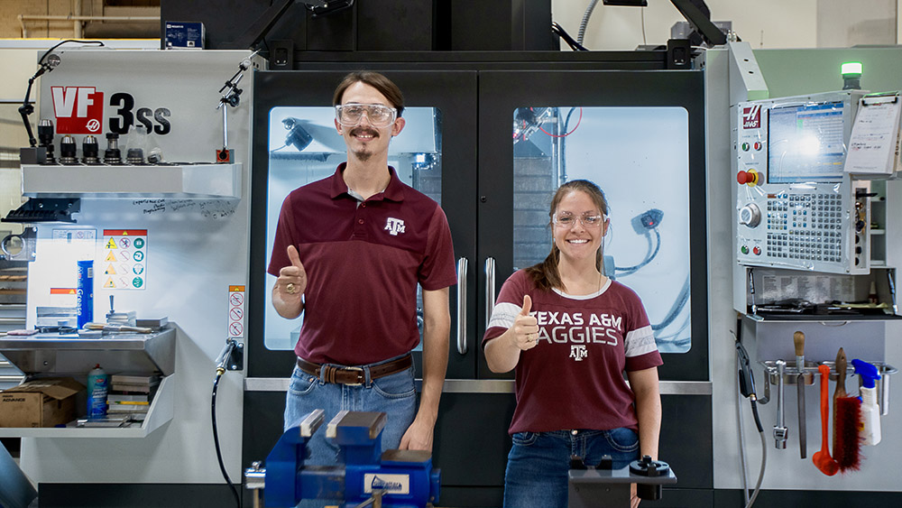 Two students wearing safety glasses smile and give thumbs up while in a machining laboratory.