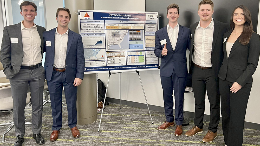 Five students in professional attire are smiling while presenting their research.