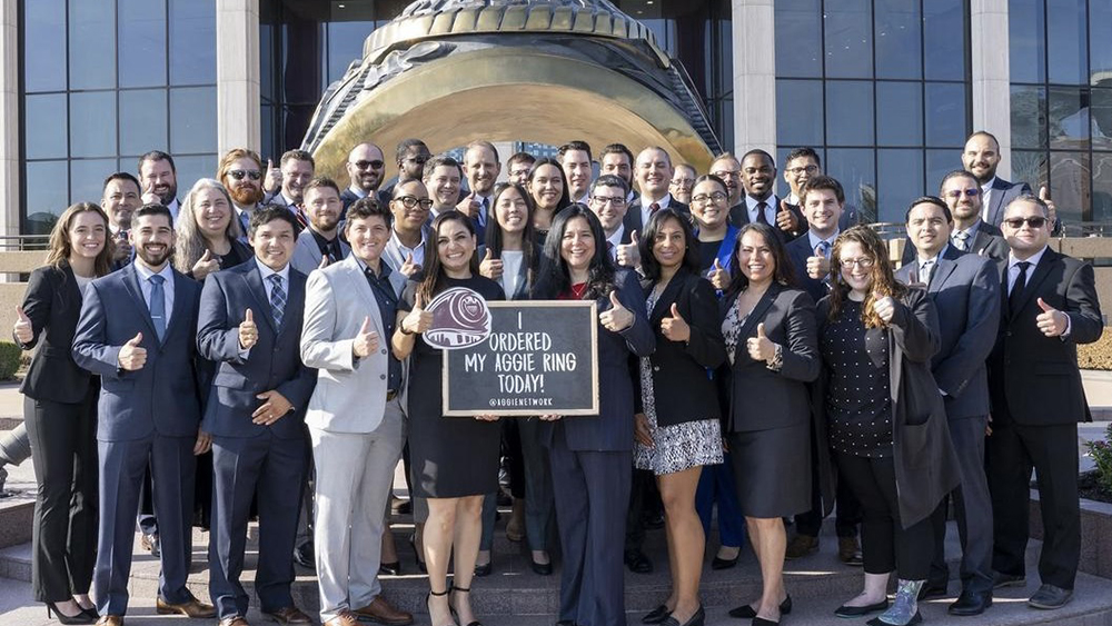 A group of individuals standing on stairs in front of an oversized Aggie ring.