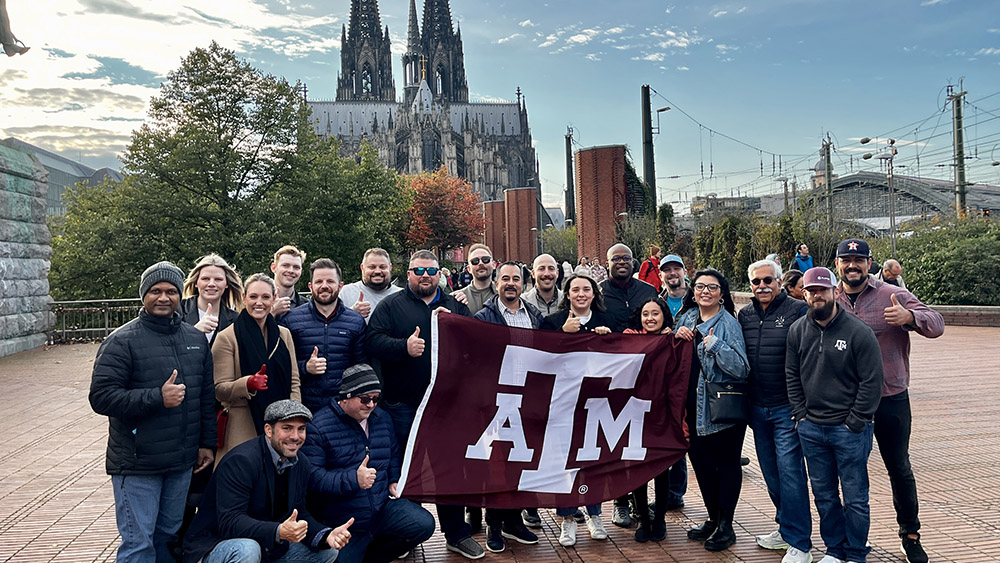 A group of individuals give thumbs up while holding a Texas A&amp;M flag.
