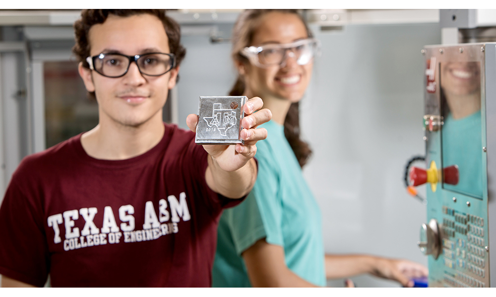 Two students wearing safety glasses are working in a lab. One is holding up a square block with the Texas A&amp;M logo inside an outline of Texas.