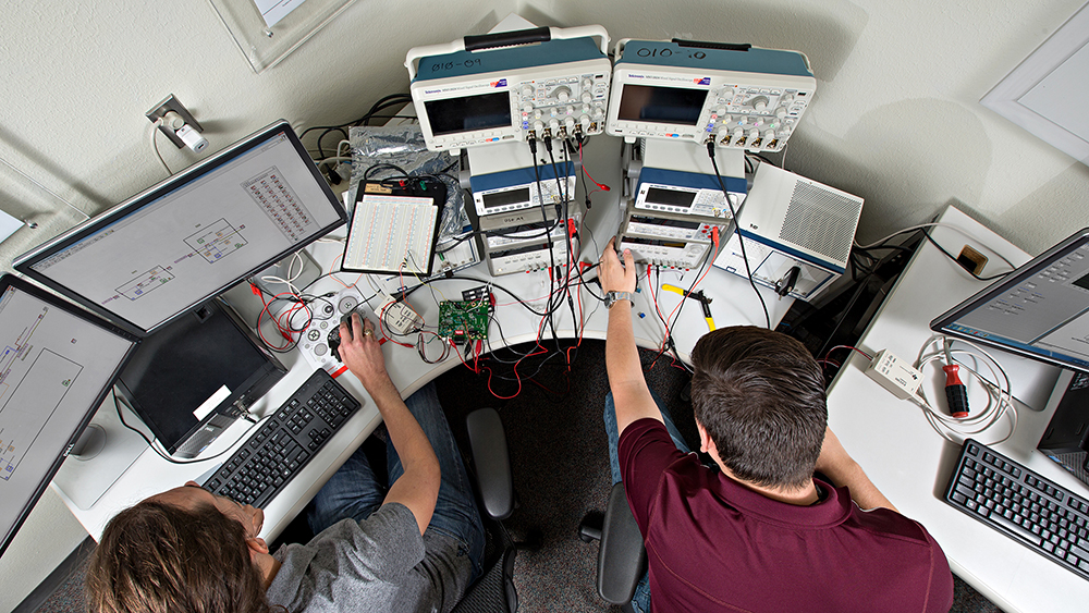 Students work at a desk filled with electronic equipment.