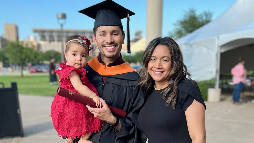 Photo of a hispanic male in graduation cap and gown holding baby while standing next to hispanic female