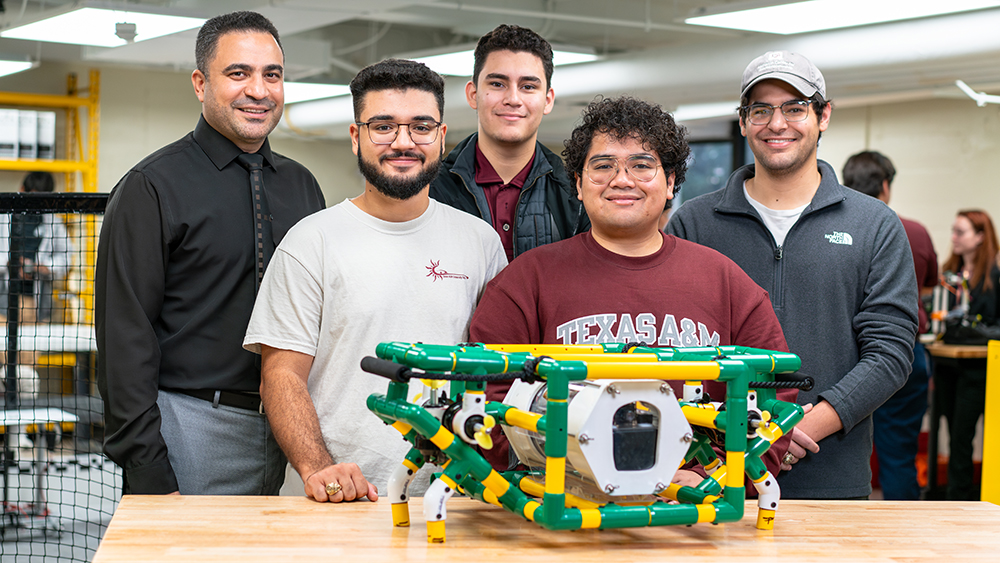 A faculty member and five students stand behind a table supporting a device constructed of plastic tubing and electronics. 