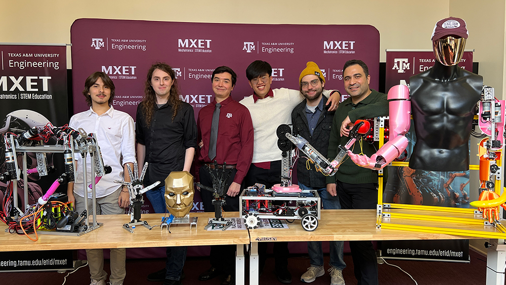 Six people stand behind a table holding robotic projects and in front of a banner with the Texas A&amp;M Engineering logo, "MXET" and "Mechatronics STEM Education."