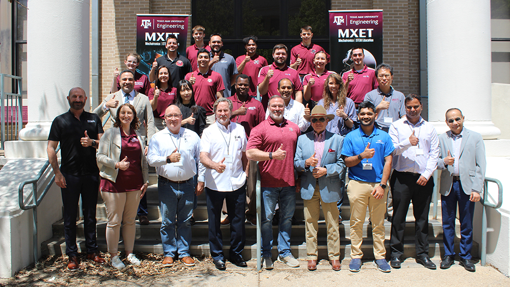 Twenty-six people standing outdoors on the steps of a building with signs behind them featuring the Texas A&M University Engineering logo and the text: MXET, Mechatronics, STEM Education.