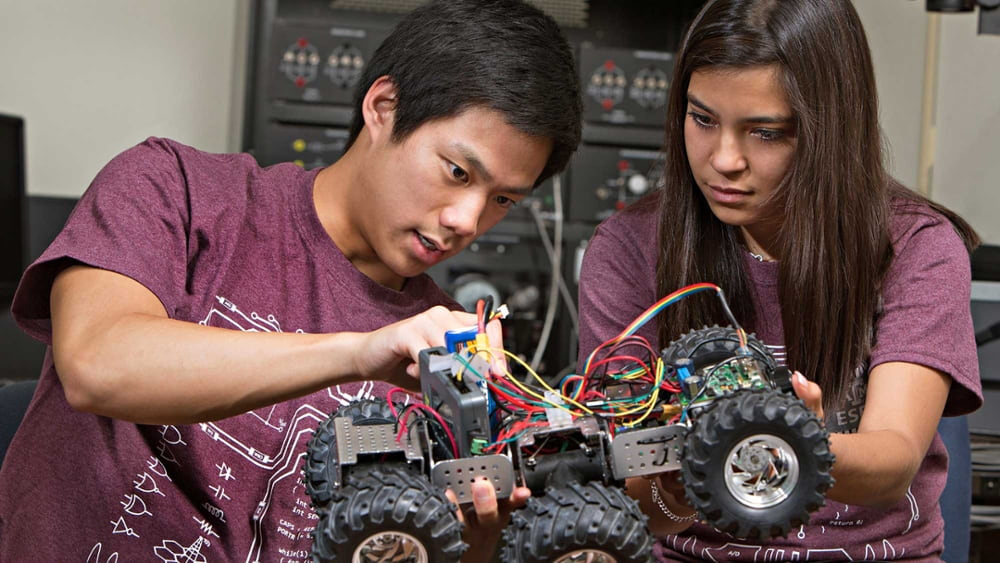 Two students hold a shoebox-sized six-wheeled robot while one student points out a wiring connection to the other student.