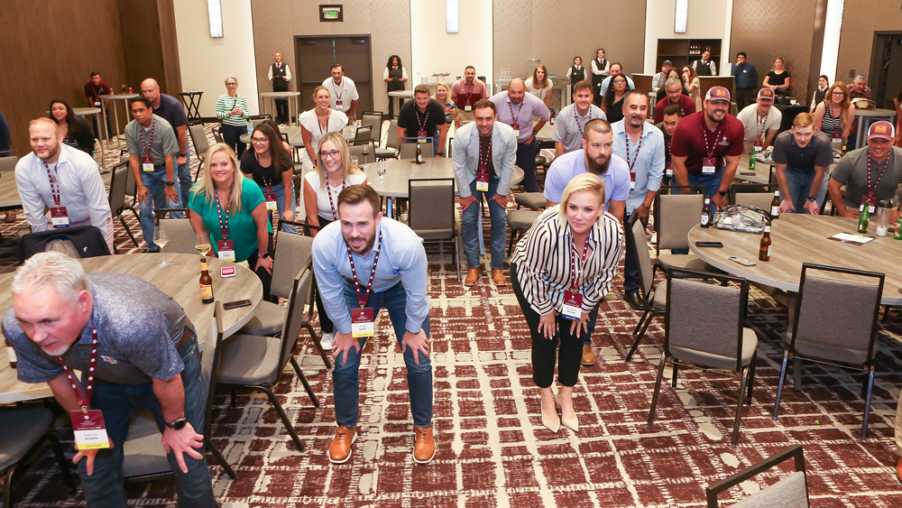 Numerous people in a large meeting space are standing partially crouched over with their hands on their knees while participating in a lecture activity