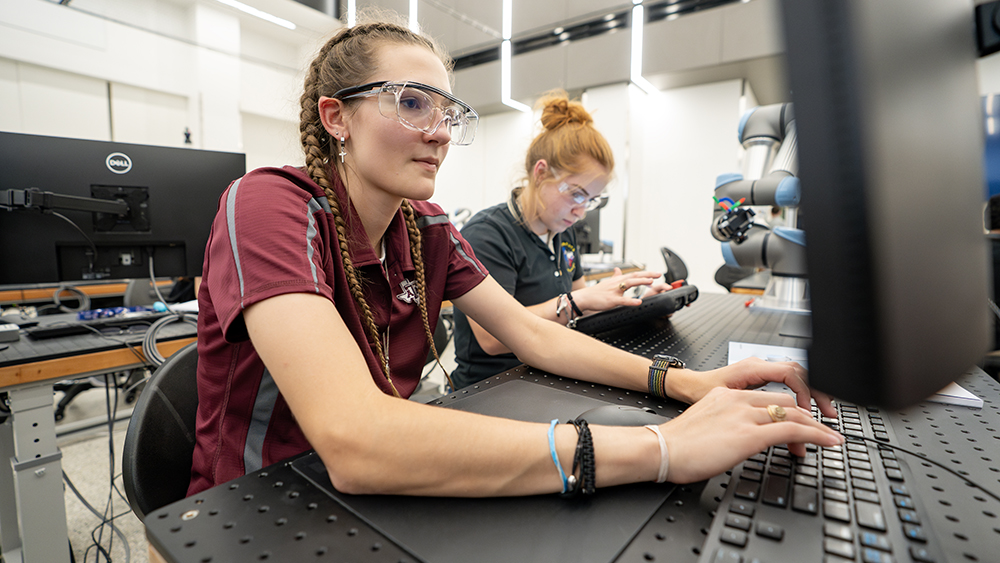 Two individuals wearing safety glasses work on computers in a robotics lab.