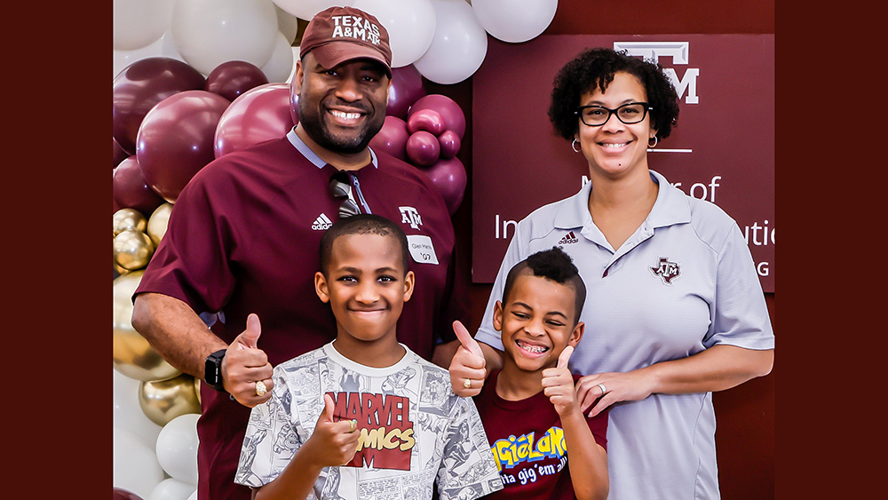 A dad, mom and two young children smile while posing in front of a backboard and balloons. Their clothing features Marvel Comics and Texas A&amp;M University logos.