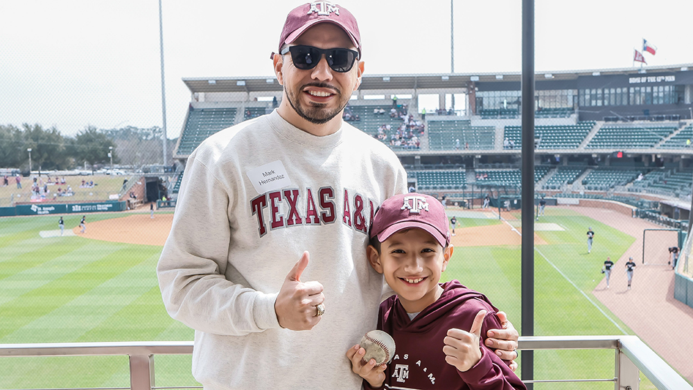 A man and a young boy holding a baseball smile at a camera as a baseball game plays on the field behind them. Logos on their clothing relate to Texas A&amp;M University.
