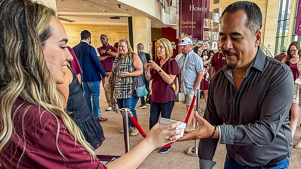 A man receives a small box from a woman at a table in a large space filled with other people waiting in lines.