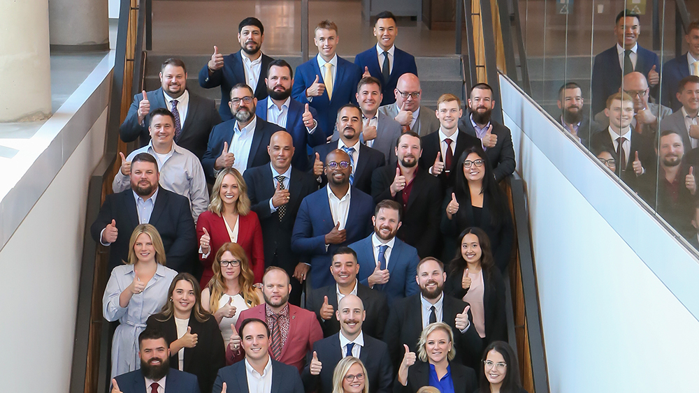 A large group of people dressed in business attire is smiling and giving thumbs-up while standing on a wide staircase.