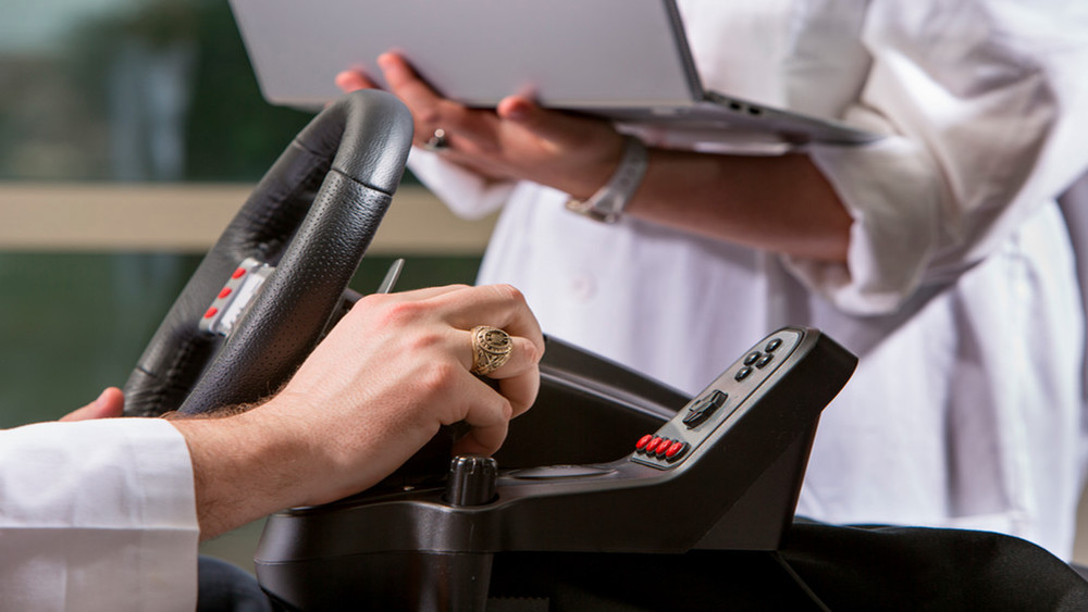 A close-up of a person's hand gripping a steering wheel, performing a driving demonstration. In the background, another person is holding a laptop.
