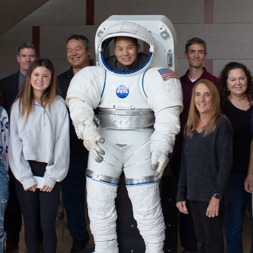 A group of NASA representatives, their families and Texas A&amp;M faculty stand together around a life-sized space suit model and smile at the camera.