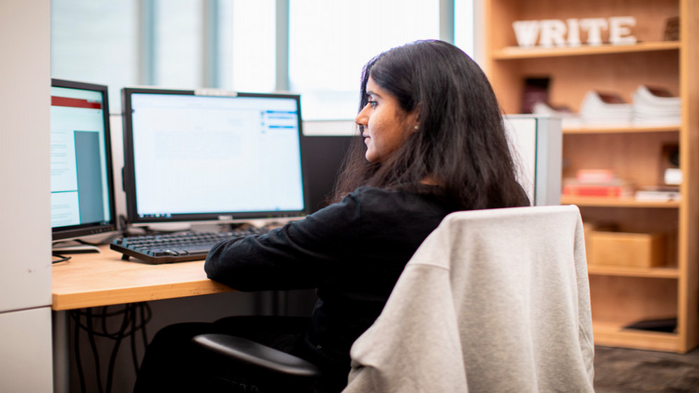 A woman with dark hair sits at a desk working on a computer. The workspace includes two monitors and a bookshelf in the back with the word "WRITE" visible.