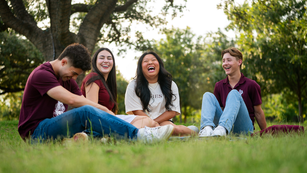 Four students sit on the grass in a park, smiling and laughing together. Three wear maroon shirts, one wears white. Trees and greenery fill the background.