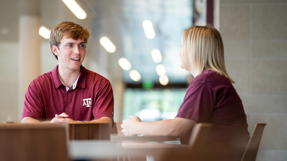 Two people in maroon shirts sit at a table, talking with each other. The background is softly blurred with lights and windows.