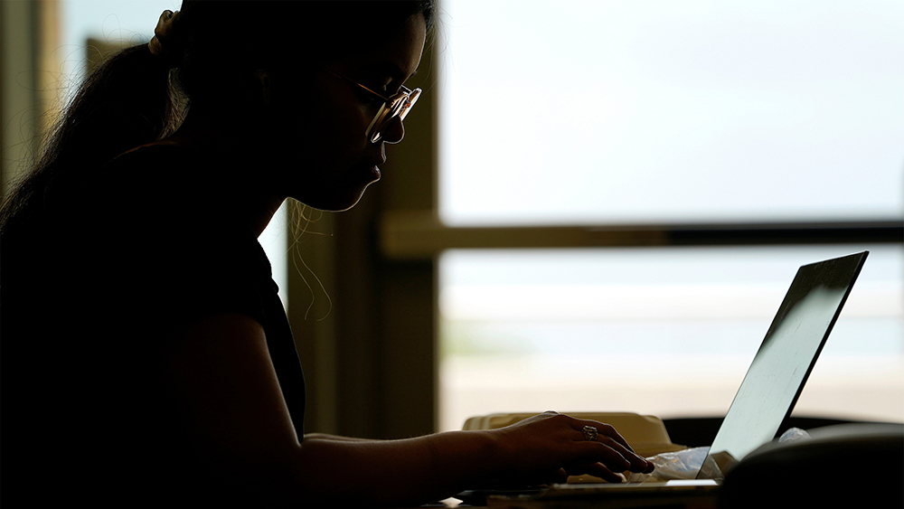 Silhouette of a student working on a laptop in front of a brightly lit window.