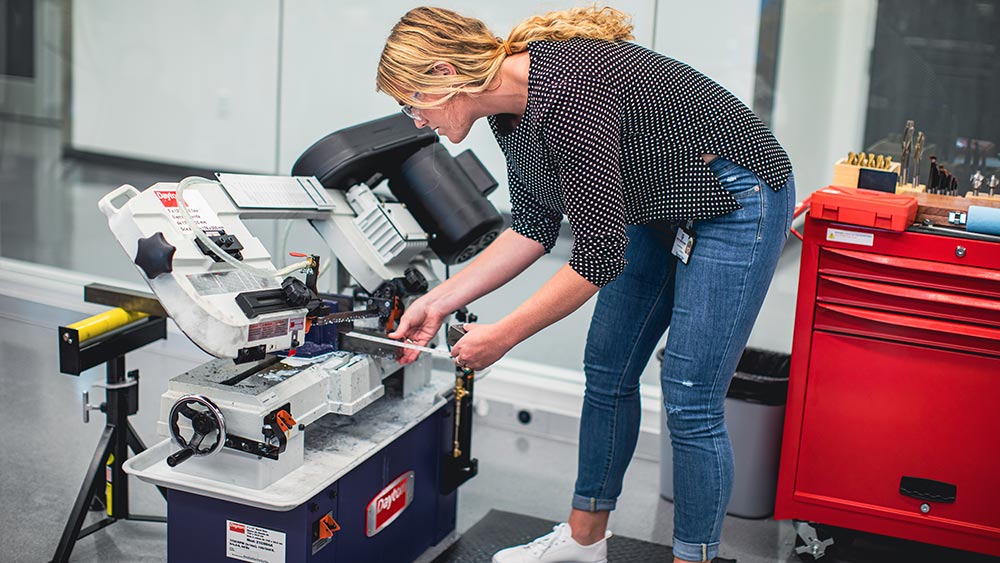 A young woman in jeans, a blouse, and safety googles is bending over a piece of heavy machinery cutting metal. 