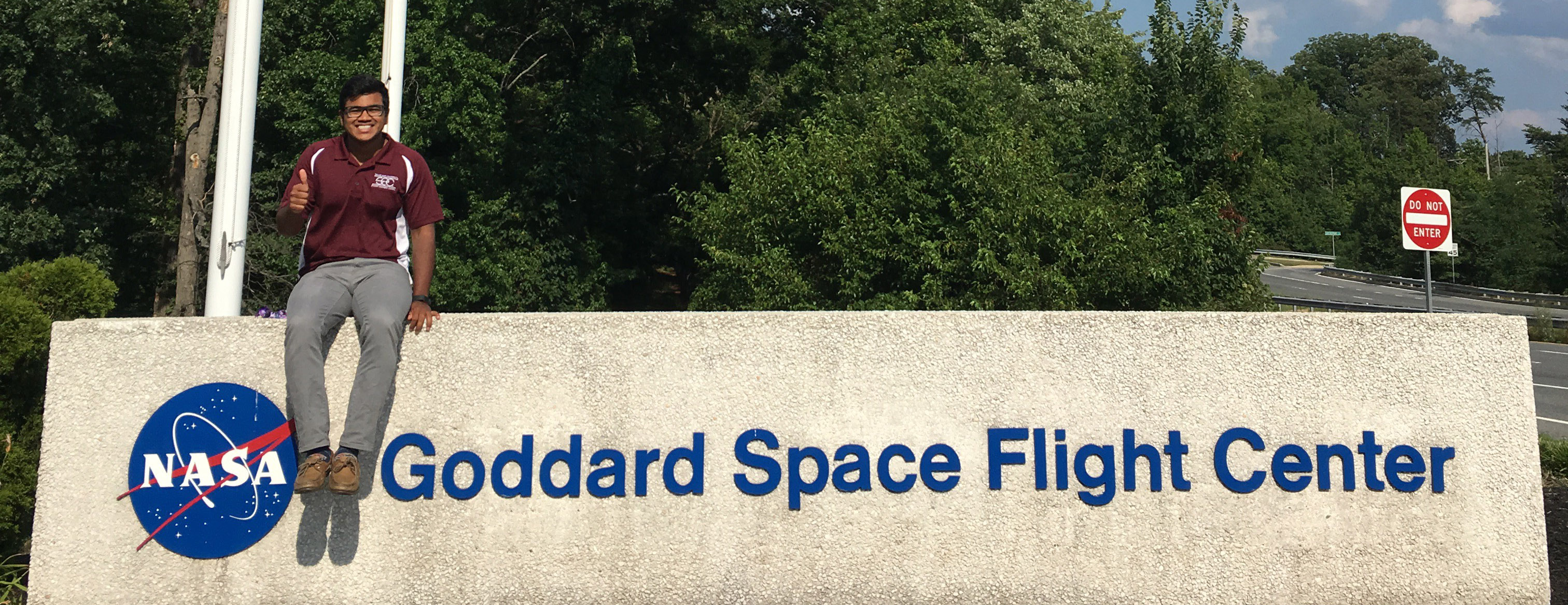 Michayal Mathew wearing maroon shirt and grey pants sits on top of sign for NASA's Goddard Space Flight Center.