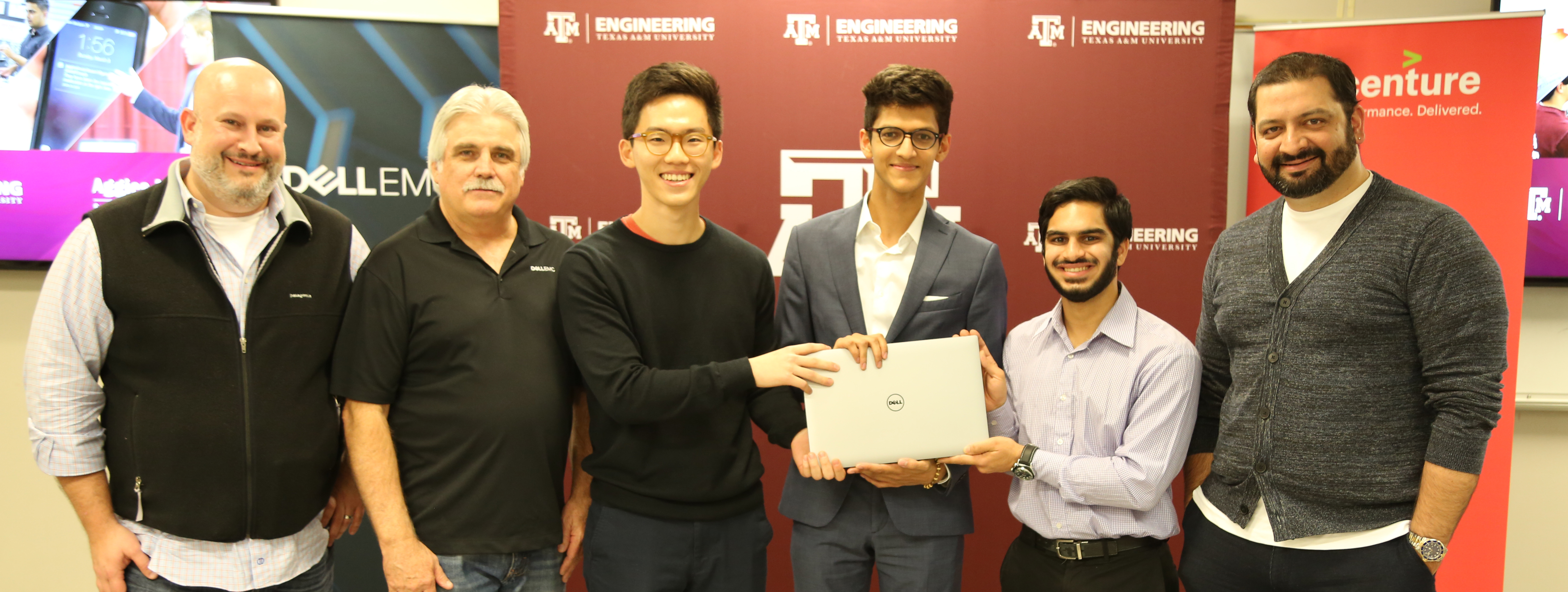 Six men (three are students holding a Dell laptop in front of them) standing in front of wall with maroon stand with the College of Engineering logo on it, a fast screen television to the left and another standing sign on the right that is red. Words are covered by man standing in front of it.