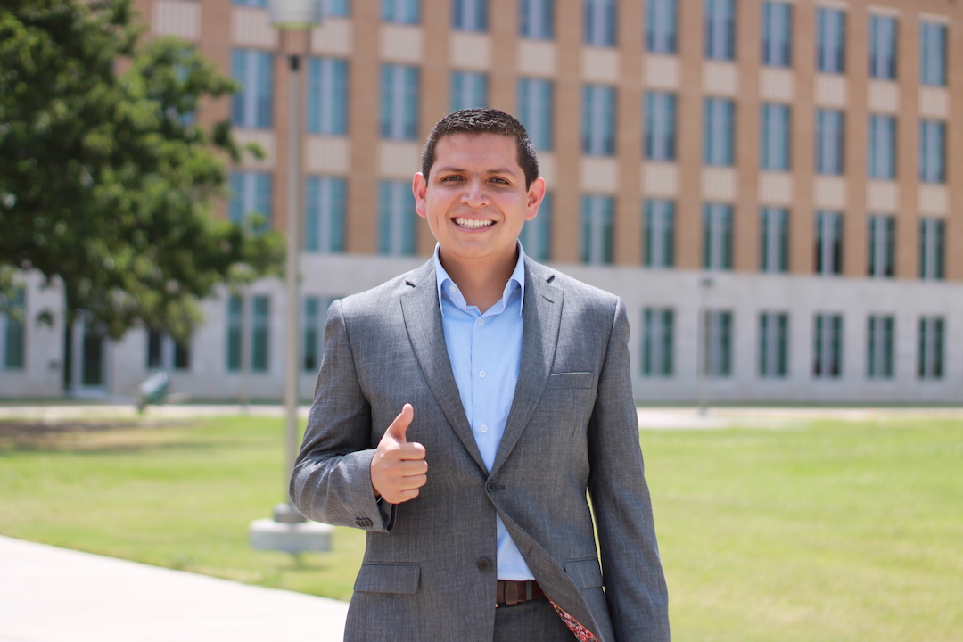 Alfredo Costilla-Reyes stands in front of building with several windows in a grey suit and blue shirt giving thumbs up with a smile.