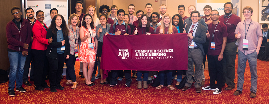 Group of computer science and engineering students and two professors with thumbs up sign. Students in the middle hold maroon banner with department name and university logo on it. They are in conference room with red carpet with yellow polka dots.