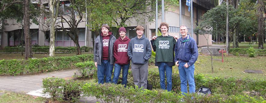 Five men (four students and one professor) standing outside in lush courtyard with lots of green bushes and trees outside of a building. The building has a lot of window. Everyone is wearing a jacket.