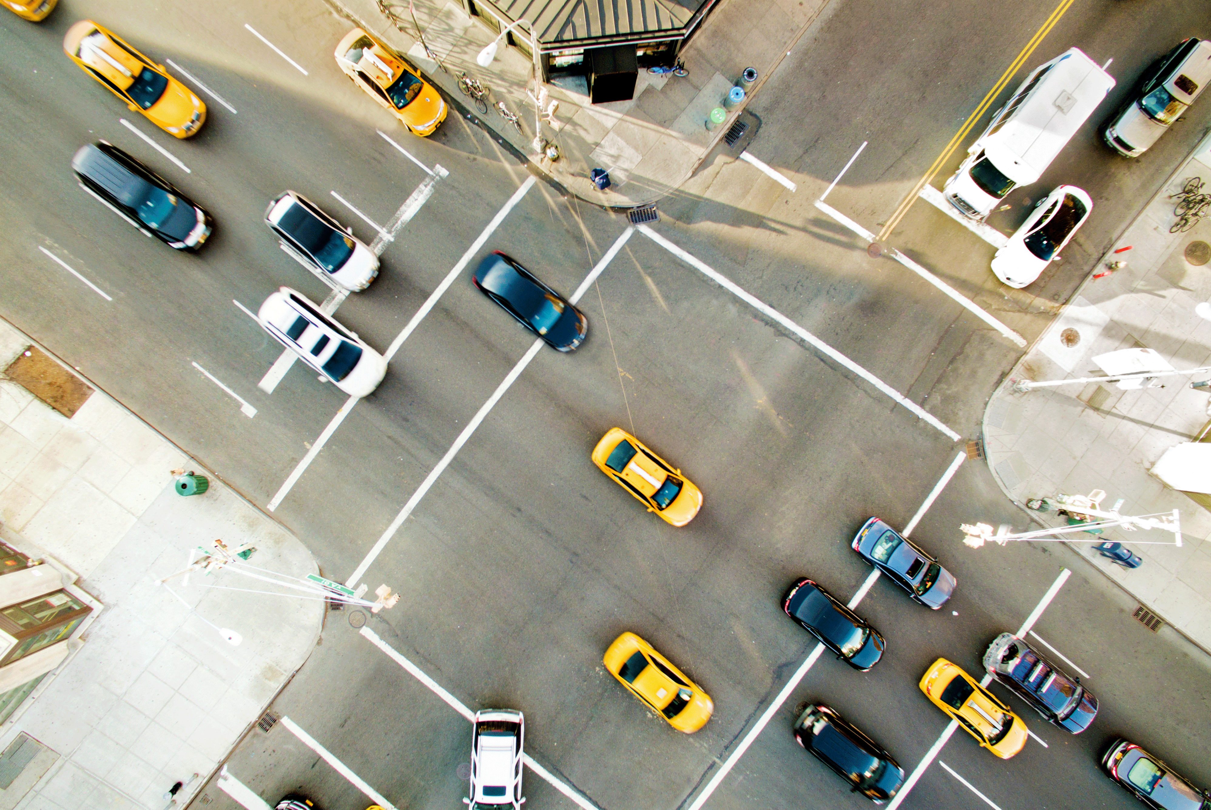 View of four way stop with traffic from above. There are white, black and yellow cars. There are traffic lights on two of the corners. Green street signs are on traffic light poles.