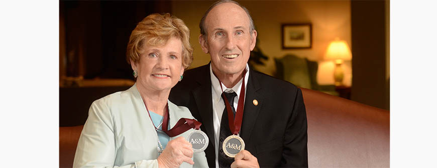 Older husband and wife in business attire with "A&amp;M" medal around their necks.