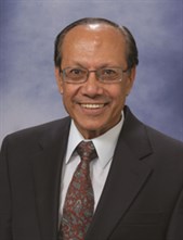 Man in formal headshot. He is wearing a black suit jacket, white collared shirt and a maroon tie with a design on it. He is wearing glasses.