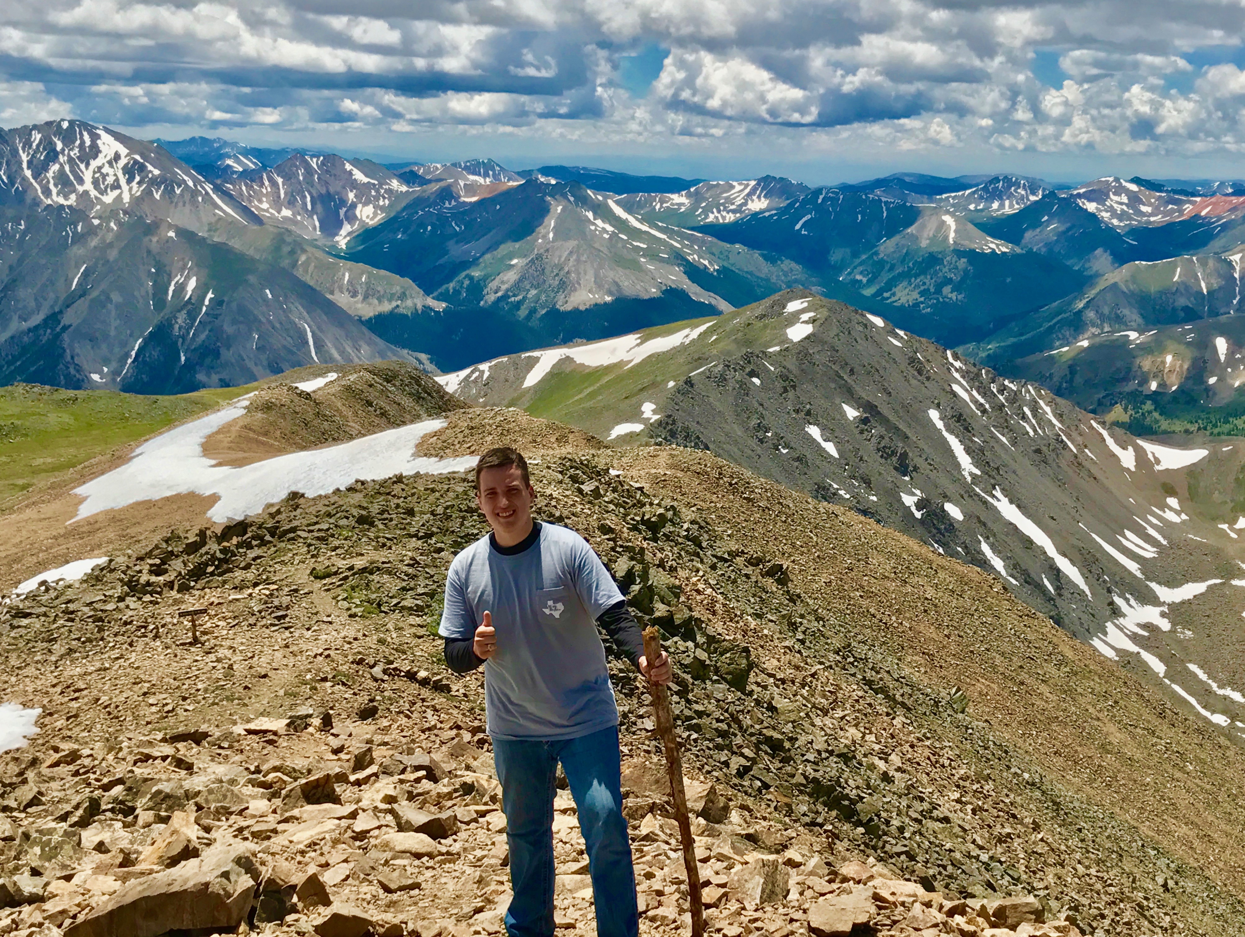Young male student standing on top of mountain holding a walking stick and giving thumbs up. Behind him is a mountain range. A little bit of snow can be seen, but the terrain is mostly grass, brown dirt and rocky.