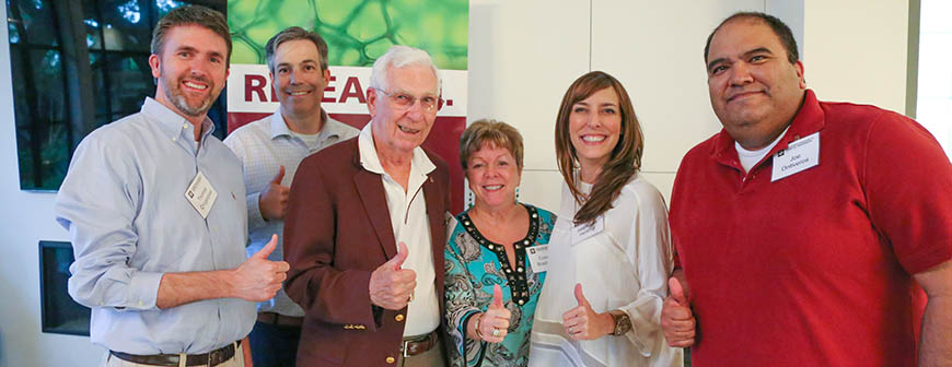 Group of four men and two women giving thumbs up to camera. Man in the middle is wearing maroon suit jacket.
