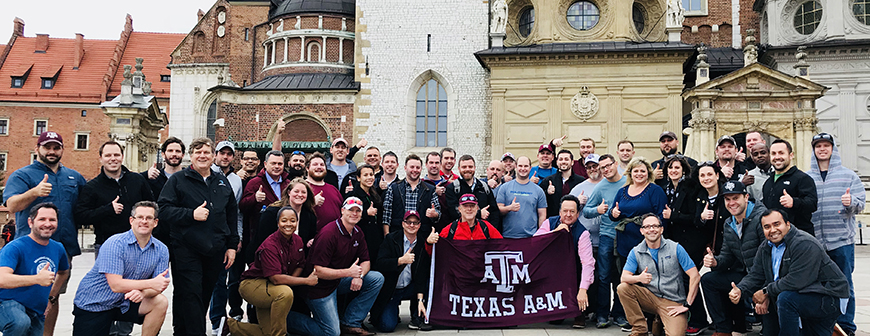 Large group of students and professors in front of old brick buildings. The men in the front are holding a maroon Texas A&amp;M flag.