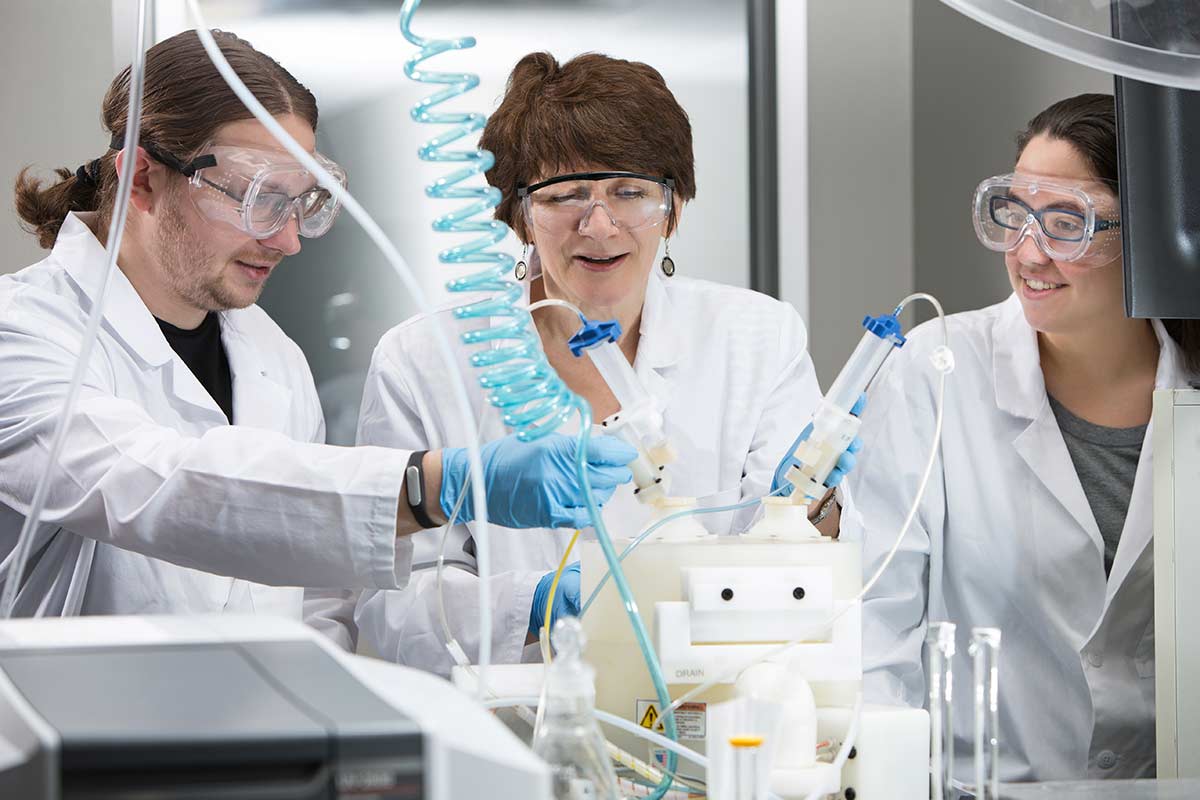 Two women and one man in lab. They are all wearing a white lab coat, goggles and blue, plastic gloves.