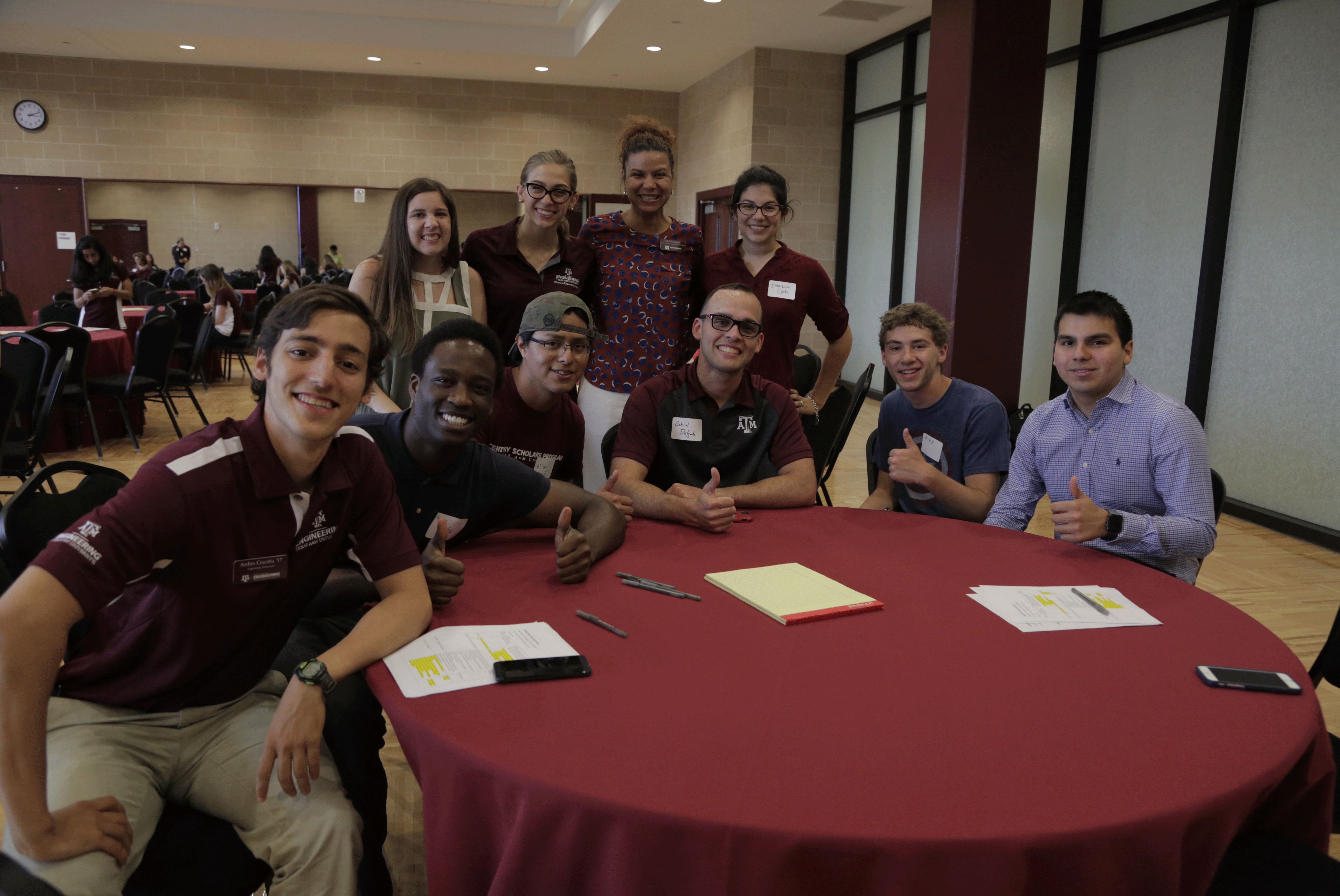 Group of six smiling male students sitting at round table with maroon table cloth. All are giving a thumbs up to the camera. Four women in business casual clothes are standing behind them smiling.