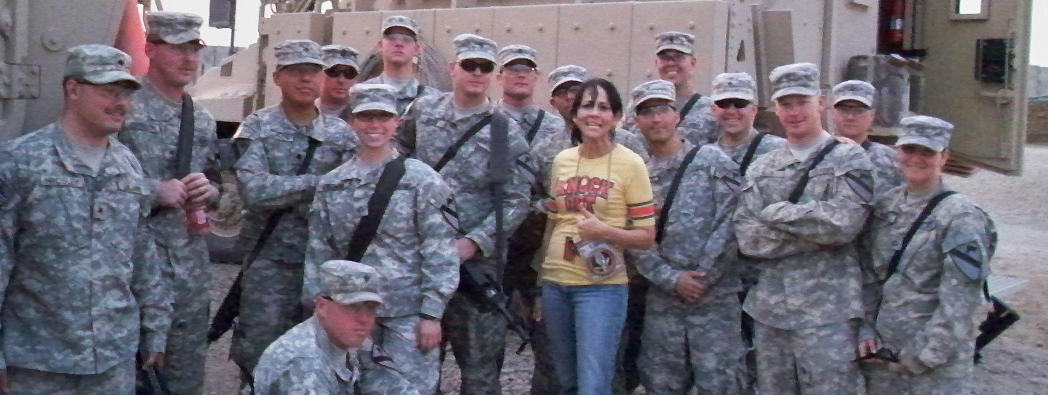 Woman in yellow shirt and jeans with group of soldiers in camouflage fatigues and hat. 