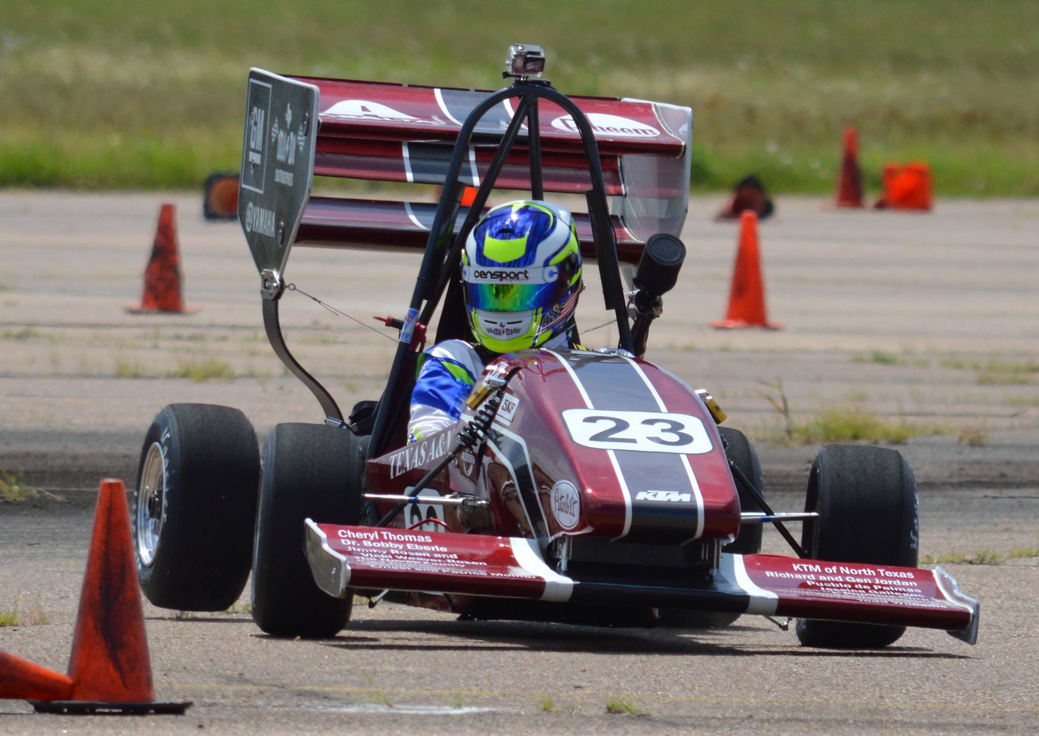 Person in blue, white and neon green racing suit and helmet in maroon, black and white race car. Number on front of car is 23.
