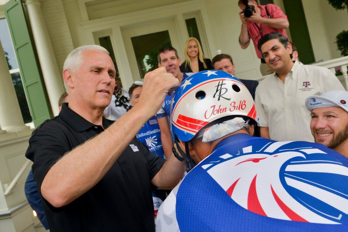 Older caucasian man with white hair in a black shirt signs helmet of bike rider. Helmet is red, blue and silver with a patriotic design. Small crowd of people are standing behind him.