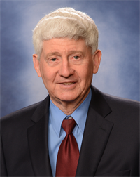 Man with white hair wearing a black blazer, light blue collared shirt and maroon tie. The backdrop is a dark blue with a bit of white. It is a formal headshot.