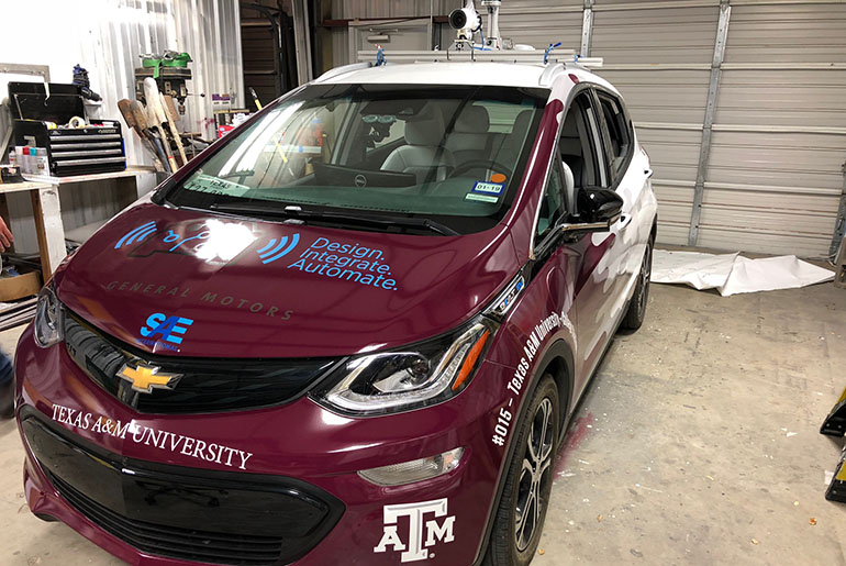 Maroon and white car sitting in garage. The car has many stickers naming different companies on it. There is a white camera attached to the top.