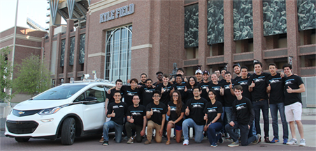 Large group of students in black T-shirts standing (the front is bent down on one knee) next to white car in front of brick exterior of Kyle Field football stadium.