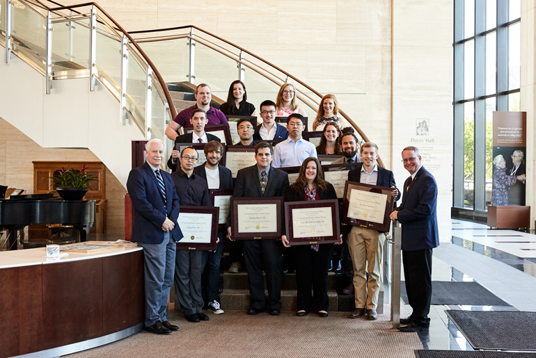 Group of students holding framed certificate posing at the bottom of a stair case. Two professors (one on each side) are standing in front of them.