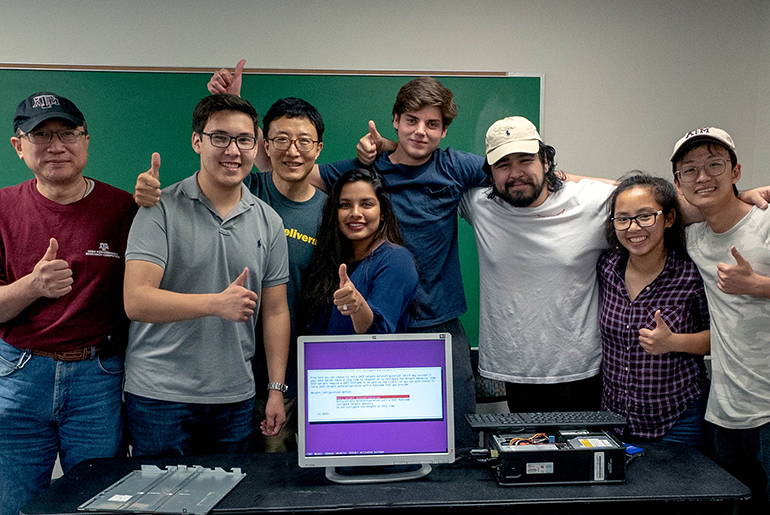 Group of students (2 female and 5 males) with male professor wearing casual clothing (three men are wearing baseball hats) in classroom standing behind a table with a computer and hard drive. The computer has a purple screen and white box displayed. There is a green chalkboard behind them.