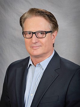 Man with short brown hair and glasses shown from the shoulders up in a formal headshot. He is wearing a dark blue/black blazer and blue and white striped shirt.