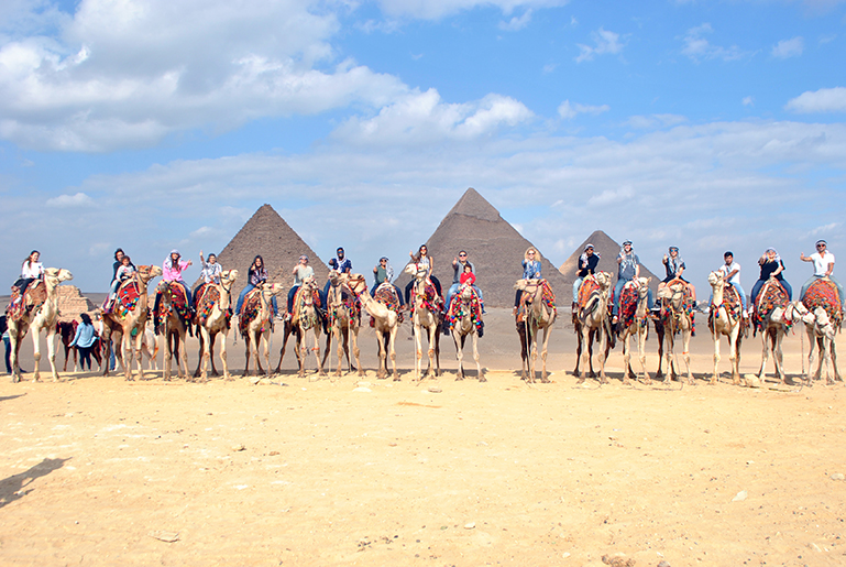Group of seventeen students in a desert in Egypt on camels. Three pyramids are in the behind them. All of the students are giving a thumbs up.