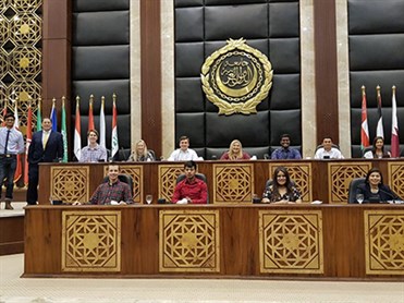 Students in a government room. There is a big, gold seal on the black paneling behind them, along with a series of different flags on flag poles. The students are sitting in 2 rows of stands. Two are standing on the left side.