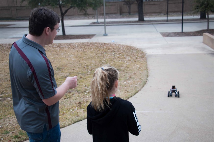 Undergraduate industrial engineering student at Texas A&M instructs fourth grade girl on steering small robot rolling down sidewalk in front of them
