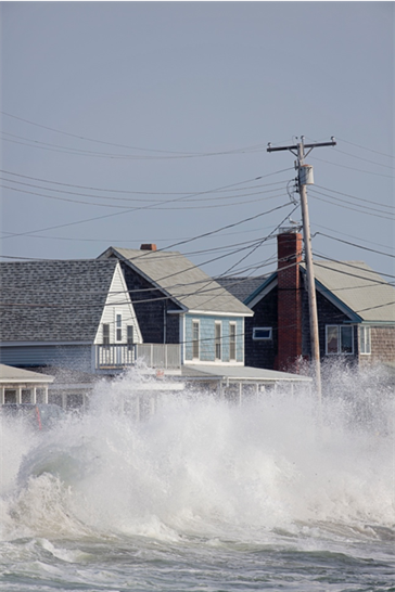 Row of houses right on the shore of a beach. There is a big wave coming towards them.